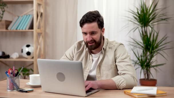 Um homem barbudo usa um laptop enquanto está sentado em uma mesa em seu escritório em casa, recebe uma mensagem com boas notícias e fica muito feliz enquanto celebra uma vitória. Fecha. Movimento lento pronto 59.97fps. — Vídeo de Stock