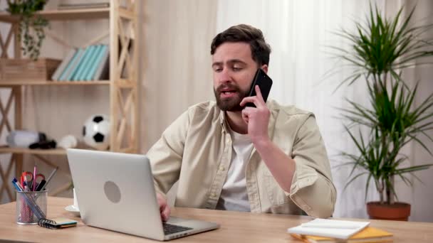Un homme s'assoit à une table et travaille sur un ordinateur portable, parlant au téléphone avec un client ou un collègue. Un homme dans un bureau à domicile Travail à distance, concept d'entreprise. Ferme là. Ralenti prêt 59.97fps. — Video