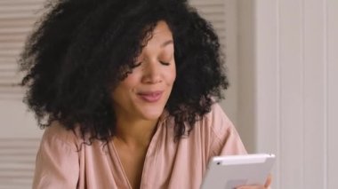 Şirin Afro-Amerikan kadın dijital tablet kullanarak bir video görüşmesinde konuşuyor. Kıvırcık saçlı siyah esmer kadın ışık odasında otururken poz veriyor. Kapatın. Ağır çekim hazır, 4K 59.97 fps 'de..