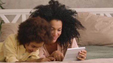 Afro-Amerikalı kadın ve küçük kız dijital tablet kullanarak görüntülü konuşma yapıyorlar. Anne ve kız poz veriyor, yatak odasında uzanmış, içi aydınlık bir şekilde. Kapatın. Yavaş çekim hazır, 4K 59.97 fps 'de.