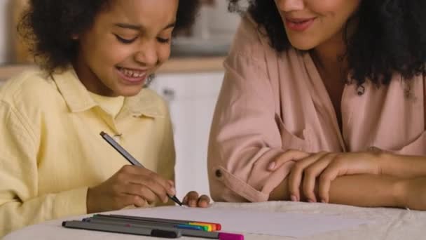 African American litle girl dessine avec des stylos feutre dans l'album. Mère et fille assises à table dans une cuisine lumineuse. Une famille heureuse passe du temps ensemble. Ferme là. Prêt au ralenti, 4K à 59,97 ips. — Video