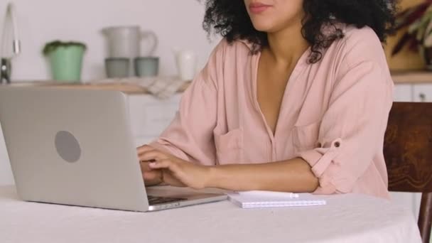 Mulher afro-americana estudando on-line, trabalho remoto usando laptop ao tomar notas no notebook. Mulher negra posando sentada à mesa na cozinha brilhante. Fecha. Movimento lento pronto, 4K a 59.97fps. — Vídeo de Stock