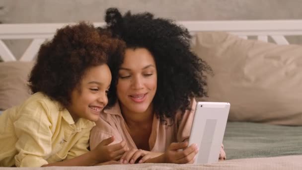 Femme afro-américaine et litle girl parler sur appel vidéo en utilisant une tablette numérique. Mère et fille posant, couchées sur le lit dans une chambre avec un intérieur lumineux. Ferme là. Prêt au ralenti, 4K à 59,97 ips — Video