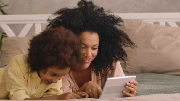 Femme afro-américaine et litle girl parler sur appel vidéo en utilisant une tablette numérique. Mère et fille posant, couchées sur le lit dans une chambre avec un intérieur lumineux. Ferme là. Prêt au ralenti, 4K à 59,97 ips — Video