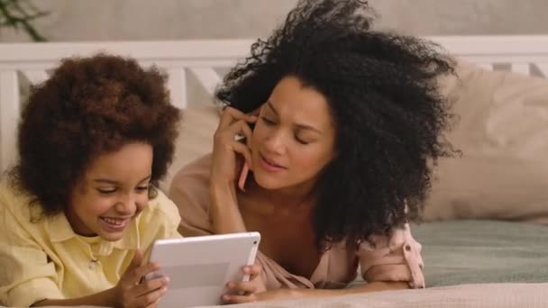 Femme afro-américaine parle sur smartphone, litle girl joue sur tablette. Mère et fille couchées sur le lit dans la chambre avec un intérieur lumineux. Ferme là. Prêt au ralenti, 4K à 59,97 ips. — Video