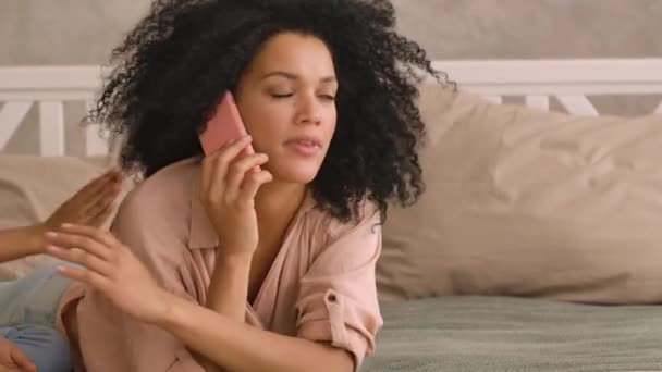 Afro-Amerikaanse vrouw praat op smartphone, klein meisje leidt haar af van het gesprek. Moeder en dochter liggen op bed in een lichte slaapkamer. Sluit maar af. Slow motion klaar, 4K bij 59,97 fps. — Stockvideo