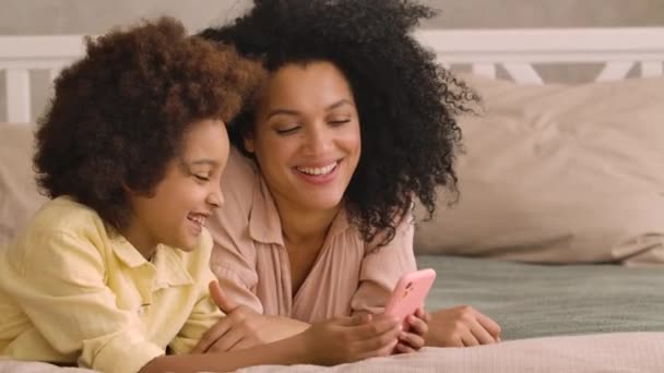Афроамериканка и маленькая девочка разговаривают по видеосвязи с помощью смартфона. Мать и дочь позируют, лежат на кровати в спальне с ярким интерьером. Закрывай. Медленное движение готово, 4К при 59,97 кадр / с. — стоковое видео