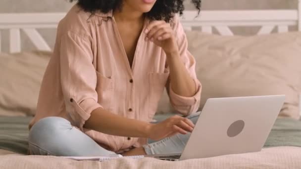 Mujer afroamericana que estudia en línea o a distancia de trabajo con el ordenador portátil, toma notas en el cuaderno. Mujer negra posando sentada en la cama en un dormitorio luminoso. De cerca. Cámara lenta lista, 4K a 59.97 fps. — Vídeo de stock