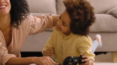 Küçük Afrikalı Amerikalı kız kablosuz kumandayla oyun oynuyor ve zafere seviniyor. Anne ve kızı aydınlık evlerinin olduğu bir odada yerde yatıyor. Kapatın. Yavaş çekim hazır, 4K 59.97 fps 'de..