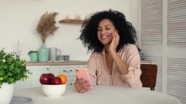 Şirin Afro-Amerikan kadın cep telefonuyla video görüşmesinde iletişim kuruyor. Kıvırcık saçlı siyah esmer kadın ışık odasında otururken poz veriyor. Ağır çekim hazır, 4K 59.97 fps 'de..