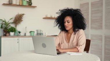 İnternette ya da uzaktan çalışan sevimli Afro-Amerikan kadın dizüstü bilgisayarla çalışırken not alıyor. Parlak mutfakta bir masada poz veren siyah kadın. Ağır çekim hazır, 4K 59.97 fps 'de..