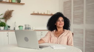 Şirin Afro-Amerikalı kadın çevrimiçi çalışıyor ya da uzaktan çalışıyor, dizüstü bilgisayar kullanıyor, yorgun ve rahatlatıcı. Parlak mutfakta masada poz veren siyah kadın. Ağır çekim hazır, 4K 59.97 fps 'de..