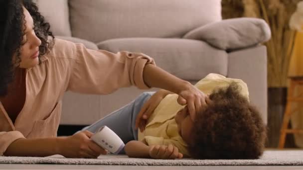 Afro-Amerikaanse vrouw meet de temperatuur van ziek klein meisje met contactloze thermometer. Moeder en dochter liggen op de vloer in de lichte huiskamer. Sluit maar af. Slow motion klaar, 4K bij 59,97 fps. — Stockvideo