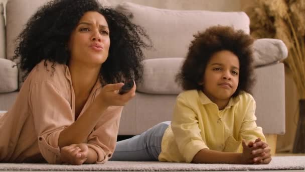 Afro-Amerikaanse vrouw en klein meisje kijken tv-kanalen met afstandsbediening. Moeder en dochter liggen op de vloer in de lichte huiskamer. Sluit maar af. Slow motion klaar, 4K bij 59,97 fps. — Stockvideo