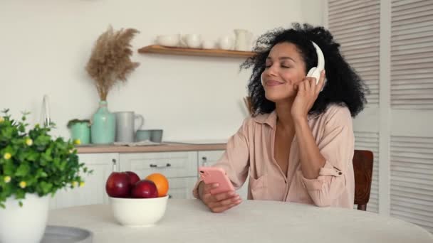 Linda mujer afroamericana en grandes auriculares blancos escucha música con placer utilizando el teléfono inteligente. Mujer negra posando mientras está sentada en la mesa de la cocina brillante. Cámara lenta lista, 4K a 59.97 fps. — Vídeos de Stock