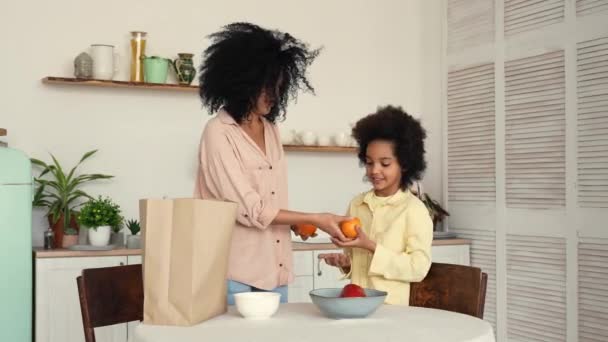 African American woman and girl taking purchases out of paper bag, putting vegetables and fruits on plates. Mother and daughter posing in bright kitchen. Slow motion ready, 4K at 59.97 fps. — Stock Video