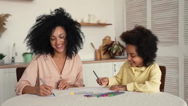 Афро-американская женщина и маленькая девочка рисуют с перьями в альбоме эскизов. Мать и дочь позируют, сидя за столом на яркой кухне. Медленное движение готово, 4K при 59,97fps. — стоковое видео