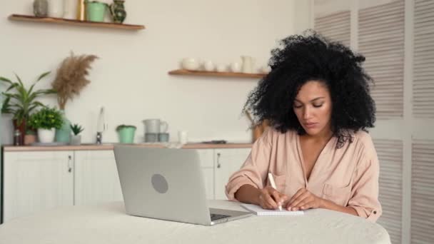 Linda mujer afroamericana que estudia en línea o a distancia trabajando con el ordenador portátil mientras toma notas en el cuaderno. Mujer negra posando sentada en una mesa en una cocina luminosa. Cámara lenta lista, 4K a 59.97 fps. — Vídeos de Stock