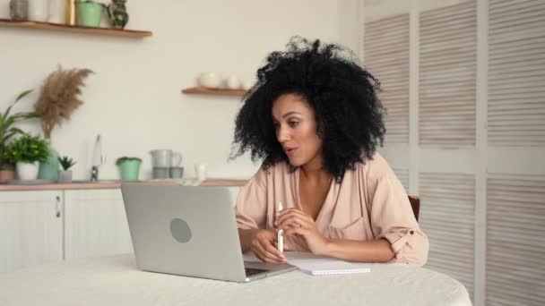 Nette Afroamerikanerin kommuniziert per Videotelefon. Schwarze brünette Frau mit lockigem Haar posiert, während sie am Tisch in der hellen Küche sitzt. Zeitlupe bereit, 4K bei 59.97fps. — Stockvideo