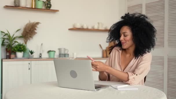 Mujer afroamericana bastante estudiando en línea o trabajando a distancia utilizando el ordenador portátil y hablando por teléfono móvil. Mujer negra posando sentada a la mesa en cocina luminosa. Cámara lenta lista, 4K a 59.97 fps. — Vídeo de stock