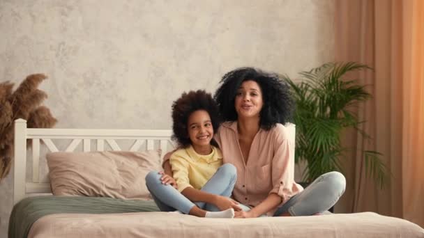 Afro-Amerikaanse vrouw en meisje knuffelen elkaar en hebben plezier met praten. Moeder en dochter poseren, zittend op het bed in de slaapkamer met helder interieur. Slow motion klaar, 4K bij 59,97 fps. — Stockvideo