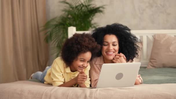 Afro-Amerikaanse vrouw en klein meisje praten op een video gesprek met een laptop. Moeder en dochter poseren, liggend op het bed in een slaapkamer met een helder interieur. Slow motion klaar, 4K bij 59,97 fps. — Stockvideo