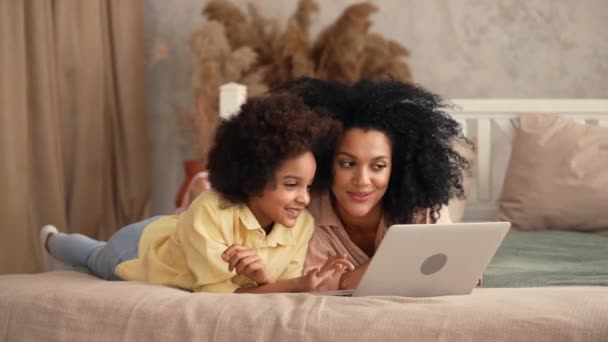 Afro-Amerikaanse vrouw en meisje typen op laptop toetsenbord, browsen inhoud en communiceren. Moeder en dochter liggen op bed in slaapkamer met helder interieur. Slow motion klaar, 4K bij 59,97 fps. — Stockvideo