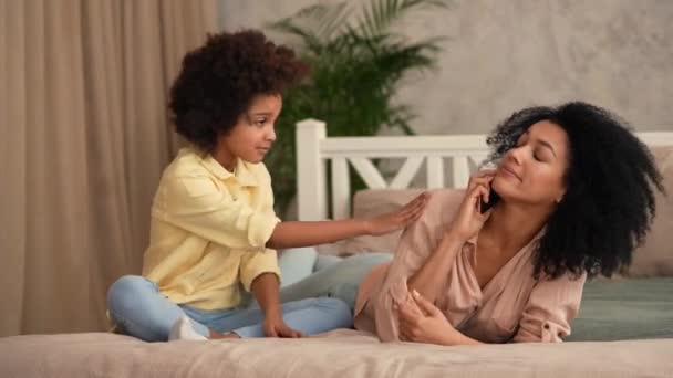 Afro-Amerikaanse vrouw praat op smartphone, terwijl een klein meisje haar afleidt van het gesprek. Moeder en dochter liggen op bed in slaapkamer met helder interieur. Slow motion klaar, 4K bij 59,97 fps. — Stockvideo