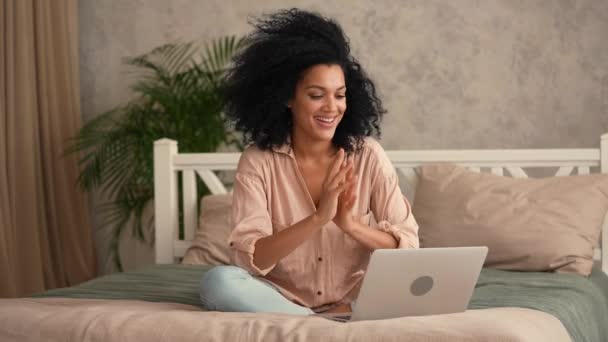 귀여운 아프리카 계 미국인 여성 이 노트북 과 웹 카메라를 사용하여 비디오 전화로 이야기하는 모습. 검은 여성 이 밝은 실내 방 이 있는 침실의 침대에 앉아 포즈를 취한다. 느린 움직임 준비 59.97fps 에서 4K. — 비디오