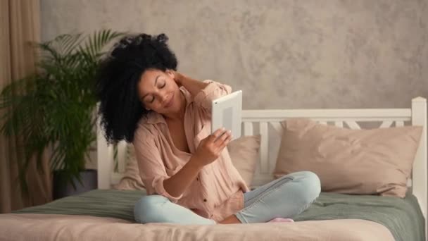 Porträtt söt afroamerikansk kvinna talar på videosamtal med digital tablett. Svart kvinna poserar sittande på sängen i ett sovrum med ljusa rum interiör. Slow motion klar, 4K vid 59,97 fps. — Stockvideo