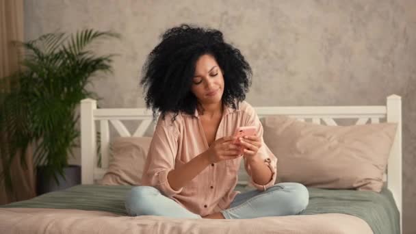 African American woman looks information on mobile phone, happily celebrates victory, received good news. Black female sitting on bed in bright bedroom. Slow motion ready, 4K at 59.97fps. — Stock Video