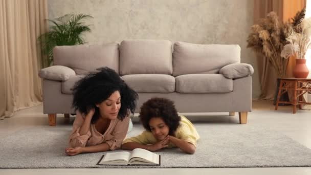 Afro American woman reads book with an interesting fairy tale to little girl. Mother and daughter posing, lying on floor in room with light home interior. Slow motion ready, 4K at 59.97 fps. — Stock Video