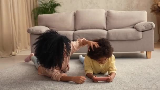 Petite fille afro-américaine joue au jeu sur smartphone et se réjouit de la victoire. Mère et fille posant, couchées sur le sol dans la chambre avec intérieur de la maison lumière. Prêt au ralenti, 4K à 59,97 ips. — Video