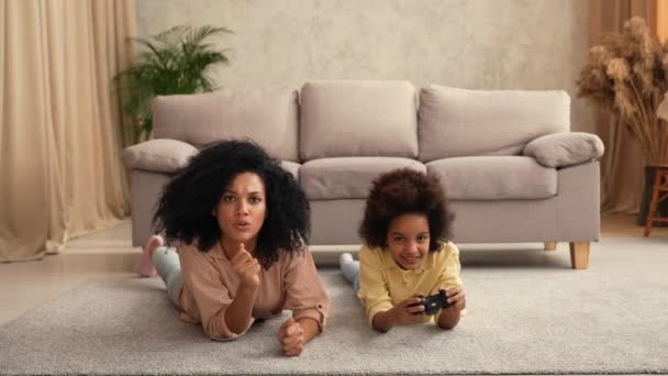Klein Afrikaans Amerikaans meisje speelt spel op draadloze joystick en verheugt zich op de overwinning. Moeder en dochter liggen op de vloer in een kamer met licht interieur. Slow motion klaar, 4K bij 59,97 fps. — Stockvideo