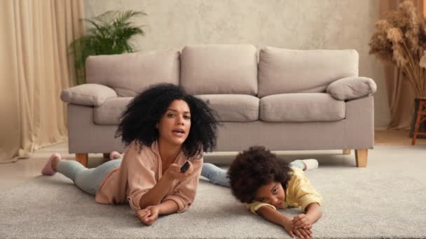 Afroameričanka a malá holka sledující dálkovým ovládáním televizi. Matka a dcera leží na podlaze ve světlém pokoji. Zpomalení připraveno, 4K při 59.97 snímcích za sekundu. — Stock video