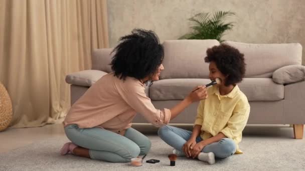 Mujer afroamericana jugando con una niña usando pinceles de maquillaje. Madre e hija posando, sentadas en el suelo en la habitación con luz interior del hogar. Cámara lenta lista, 4K a 59.97 fps. — Vídeo de stock