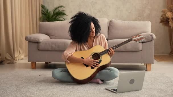 Afro-Amerikaanse vrouw speelt een akoestische gitaar, zingt liedjes en praat op videogesprek met behulp van laptop. Zwarte brunette vrouw poseren, zitten op de vloer in de lichte kamer. Slow motion klaar, 4K bij 59,97 fps. — Stockvideo