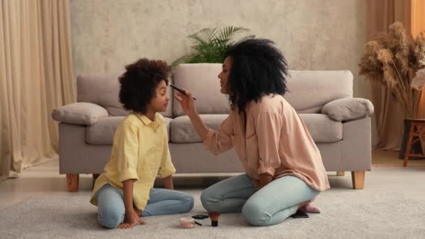 Afro-Amerikaanse vrouw spelen met kleine meisje met behulp van make-up borstels. Moeder en dochter poseren, zitten op de vloer in kamer met licht interieur. Slow motion klaar, 4K bij 59,97 fps. — Stockvideo