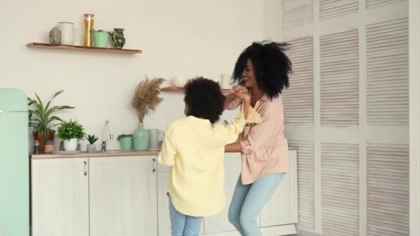 Mujer y chica afroamericanas bailando alegremente, saltando de la mano y abrazándose alegremente. Madre e hija posan en una cocina luminosa. Movimiento lento. — Vídeos de Stock