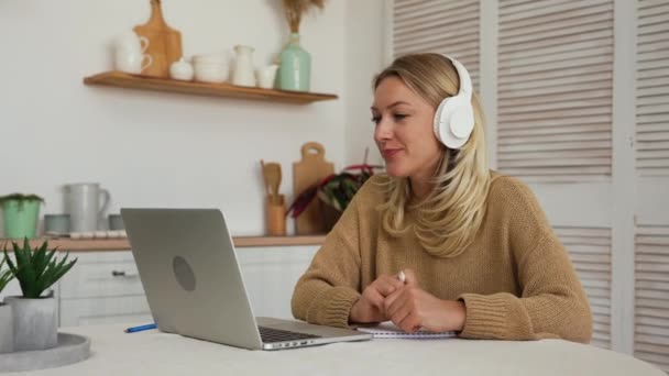Een vrouw met een koptelefoon die via een videogesprek op een laptop communiceert. Student sprekende online conferentie kantoor op afstand chat, virtuele training concept. Blond zitten aan tafel in een lichte keuken. Langzame beweging. — Stockvideo