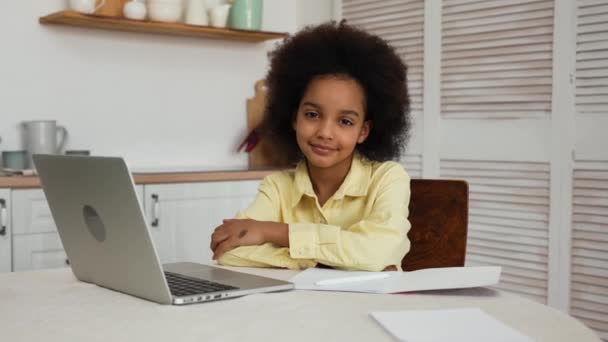 Niña afroamericana mirando a la cámara y sonriendo. Colegiala se dedica a la educación a distancia utilizando un ordenador portátil. Una adolescente está sentada en una mesa en una cocina luminosa. Movimiento lento. — Vídeos de Stock