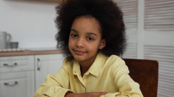 Gadis Afrika-Amerika kecil melihat ke kamera dan tersenyum. Schoolgirl terlibat dalam pembelajaran jarak jauh menggunakan laptop. Seorang gadis remaja duduk di meja di dapur yang cerah. Gerakan lambat. — Stok Video