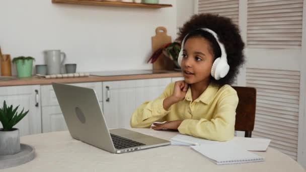 Klein Afro-Amerikaans meisje in hoofdtelefoon gebruikt laptop om video te bellen met de leraar. Online lezing, meisje steekt de hand op en beantwoordt vragen. Tiener zit aan tafel in de keuken. Langzame beweging. — Stockvideo