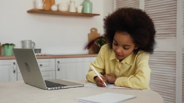 Petite fille afro-américaine utilise ordinateur portable pour appel vidéo avec le professeur. Conférence en ligne, écolière écrit des informations dans le carnet. Adolescente est assise à table dans une cuisine lumineuse. Mouvement lent. — Video