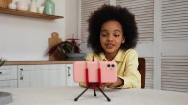 Küçük Afro-Amerikalı bir kız akıllı telefon kullanarak bir video görüşmesinde konuşuyor. Melez bir ırk kızı ilginç bilgiler ya da haberler verir. Genç kız parlak bir mutfakta bir masada oturuyor. Yavaş çekim.