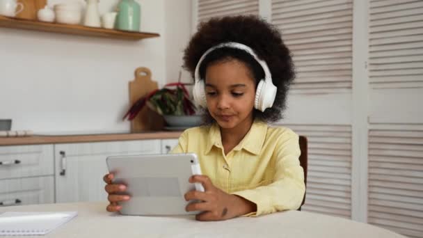 大きな白いヘッドフォンの小さなアフリカ系アメリカ人の女の子は、デジタルタブレットを使用してゲームをプレイします。オンライン学習、休憩、レジャー中に休息。10代の女の子が明るいキッチンのテーブルに座っている。スローモーション. — ストック動画