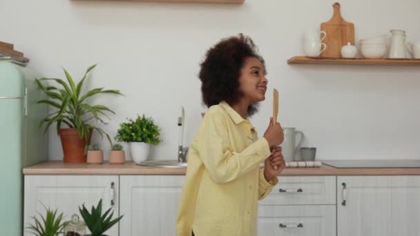 Little African American girl singing song cheerfully using kitchen spatula instead of microphone. Teenage girl posing against the background of a bright kitchen with a beautiful interior. Slow motion. — Stock Video