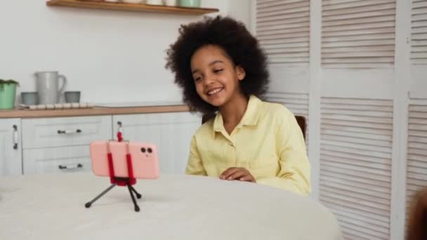 Petite afro-américaine enregistre une vidéo drôle à l'aide d'un smartphone. L'enfant noir s'amuse, fait des grimaces drôles et danse. Adolescente s'assoit à une table dans une cuisine lumineuse. Mouvement lent. — Video