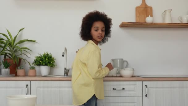 Den lilla afroamerikanska flickan lägger djupa vita skålar och muggar på bordet. Förbereder för lunch. Tonårstjej poserar mot bakgrunden av ett ljust kök med en vacker interiör. Långsamma rörelser. — Stockvideo