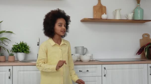 Den lilla afroamerikanska flickan lägger djupa vita skålar på bordet. Förbereder för lunch. Tonårstjej poserar mot bakgrunden av ett ljust kök med en vacker interiör. Långsamma rörelser. — Stockvideo